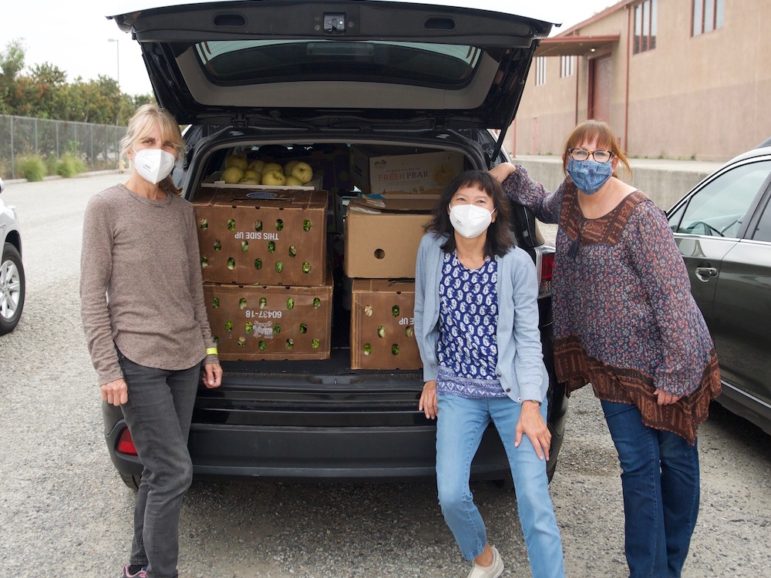 Three woman wearing face masks stand in front of a car trunk filled with produce boxes