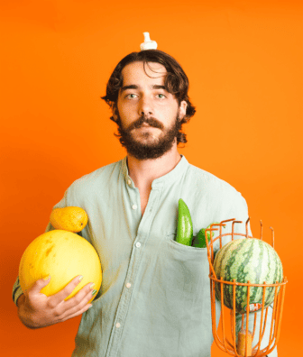 Diego balancing mushroom on head, holding melon and mango, with watermelon in fruit picker and peppers in pocket