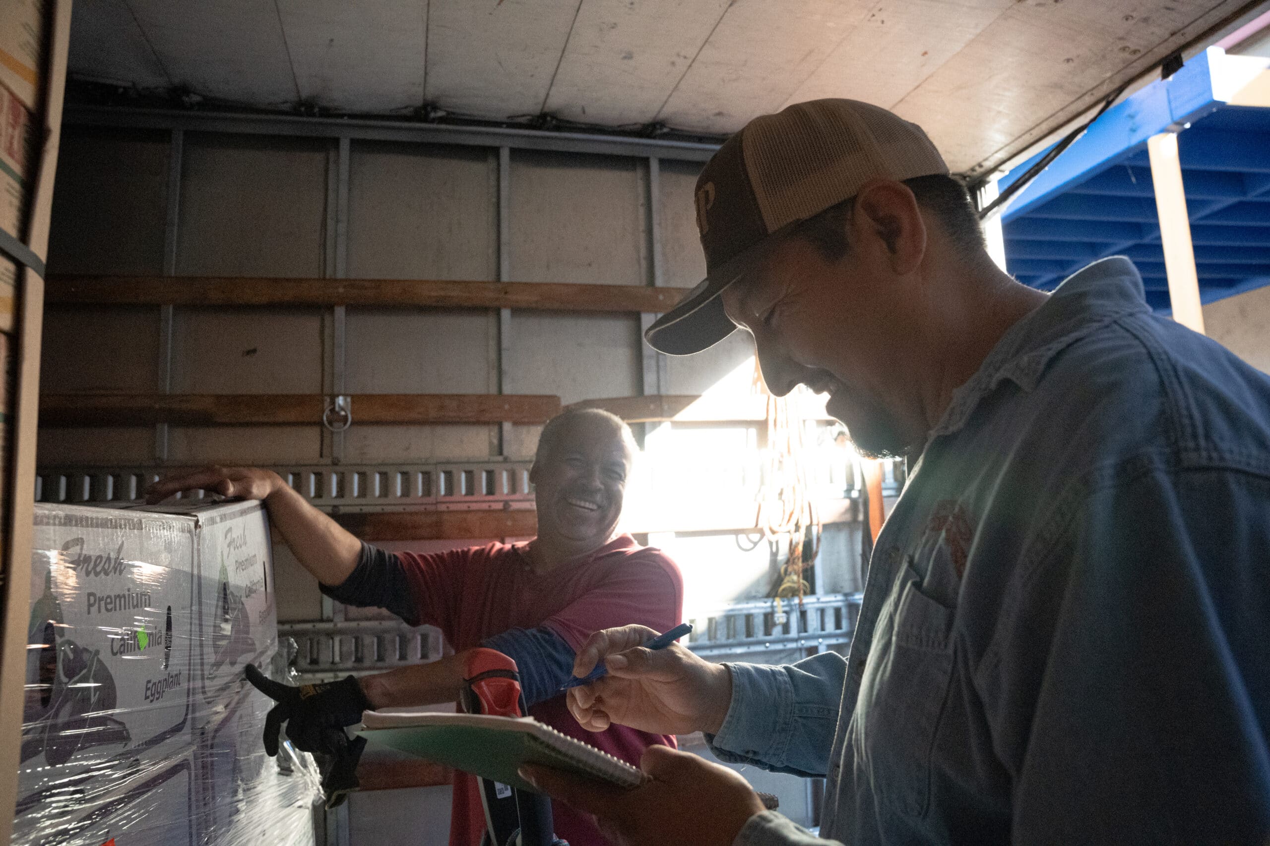 Omar smiling and pointing to boxes while Polo smiles and looks at notebook in a Food Forward truck 