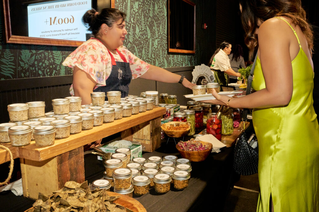 a vendor hands a jar of preserved food to a guest at a tasting table at tomorrow’s table event.